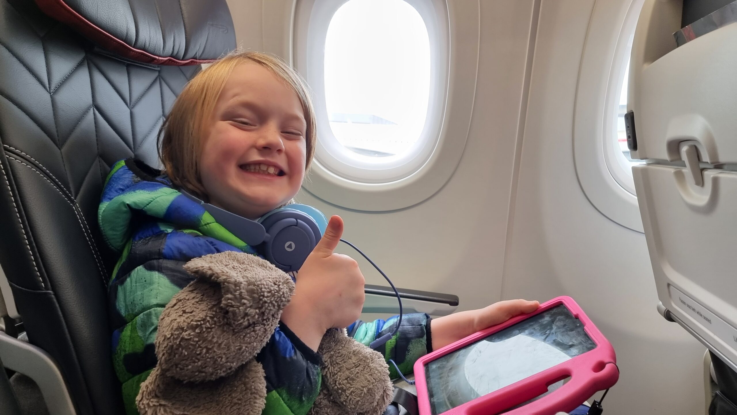 Full image of A boy smiles widely as he sits, playing with his tablet, in an aeroplane window seat.
