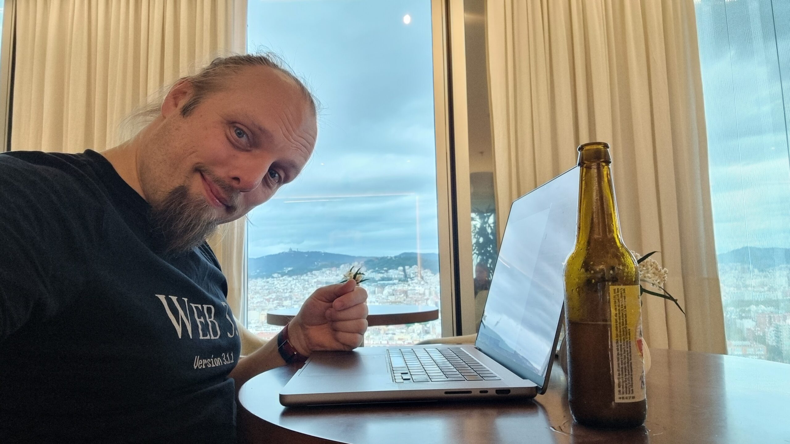 Full image of Dan sits at a laptop in a hotel bar, a view of Barcelona out of the window behind him, a beer bottle alongside him.