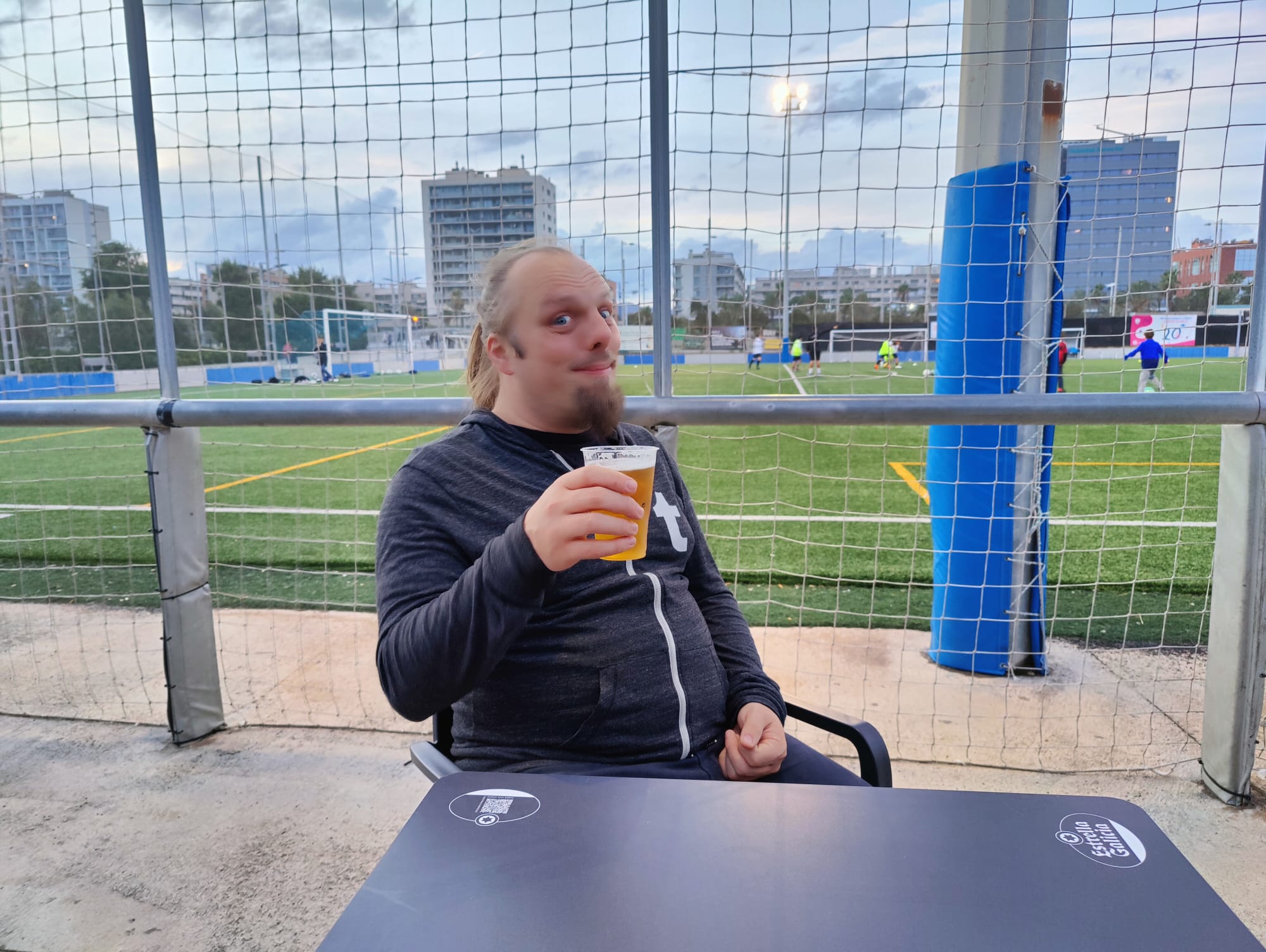 Full image of Dan drinks a beer, a children's football match is being played in the background.