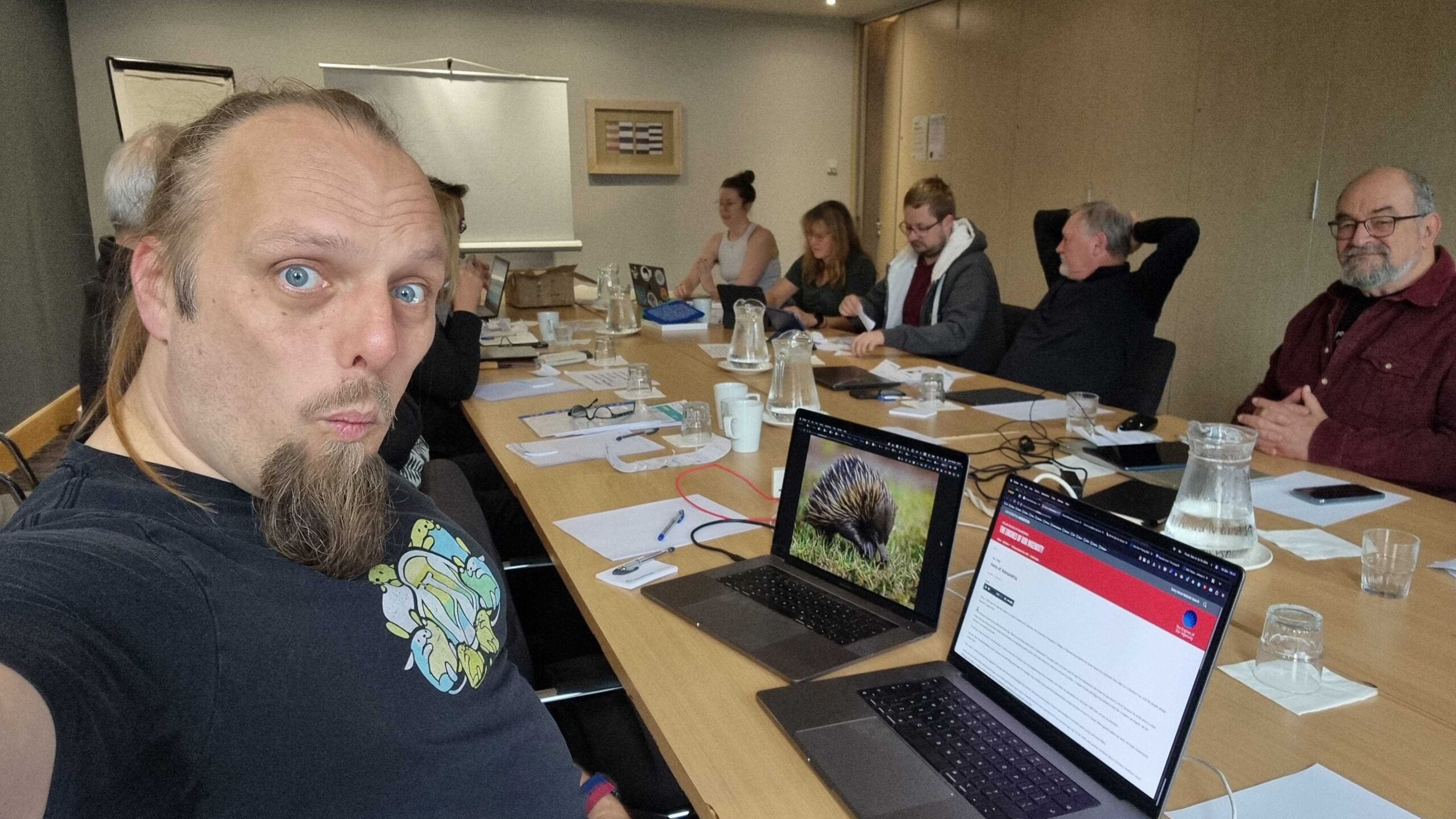 Full image of Dan sits in front of two laptops (one of which shows a photo of an echidna for some reason), in a meeting room full of casually-dressed volunteers.