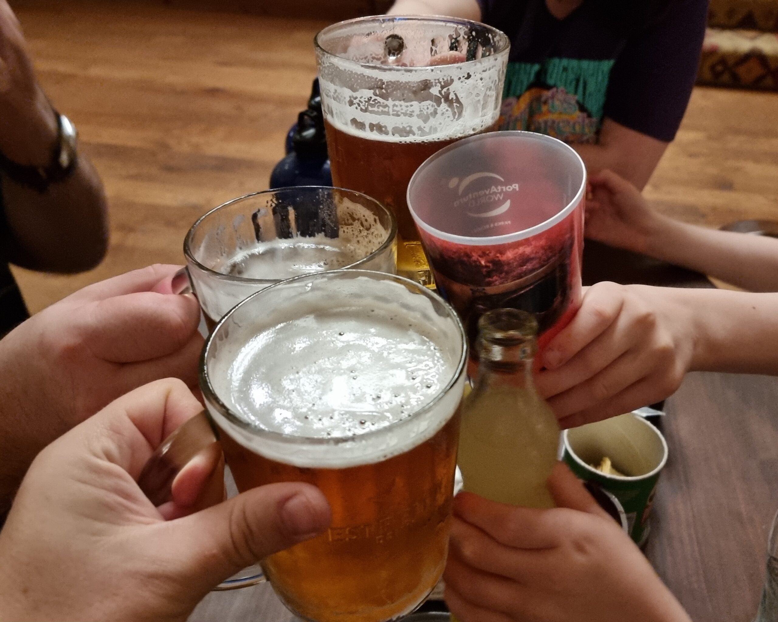 Full image of Three glasses of beer held by adult hands clink together against a glass of water and a bottle of Fanta held 6 by cold hands.