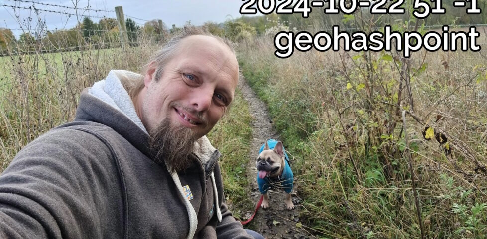 Dan Q smiles on a muddy footpath, alongside his dog, under a slightly-overcast sky.