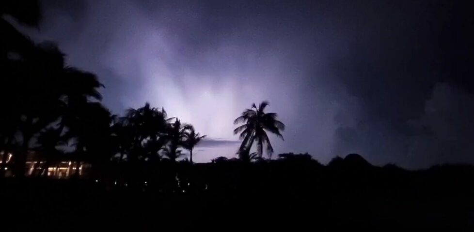 Video framegrab of sheet lightning illuminating almost the entire night sky above silhouettes of palm trees and dunes.