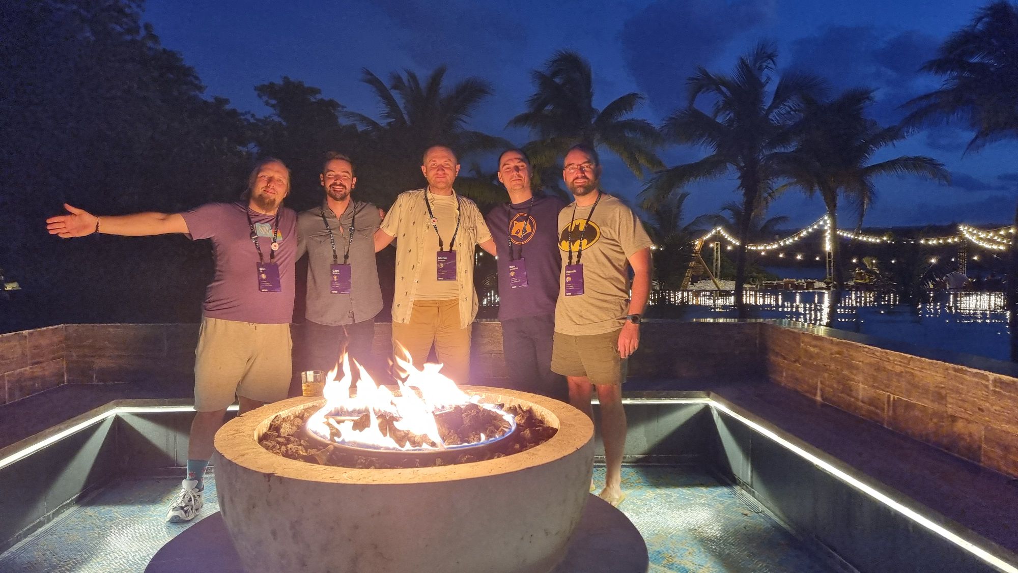Full image of Dan and four other men stand around a firepit, in front of a tropical beach and a twilight sky.