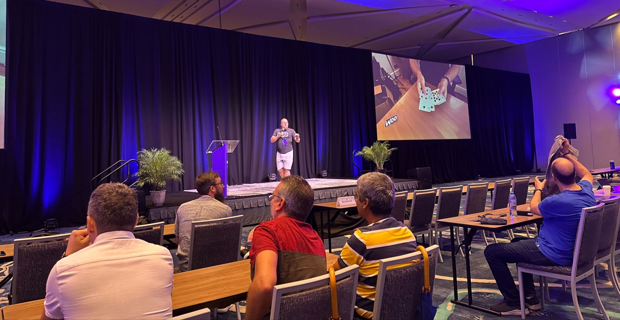Full image of Dan standing on stage in front of a seated audience; a screen behind him shows a close-up of his hands holding several playing cards.