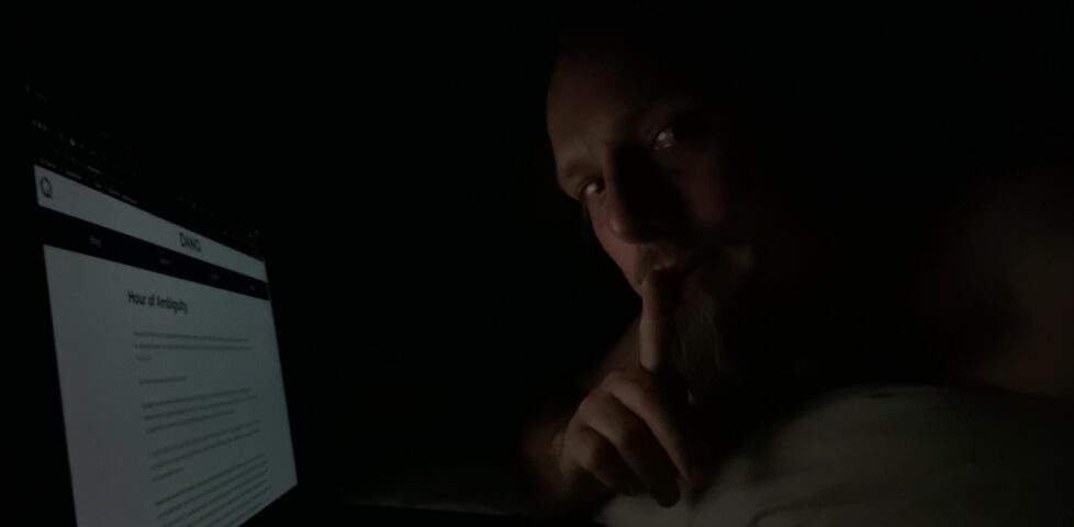Dan, lying in bed with his finger to his lips, lit by a laptop screen showing this blog post.