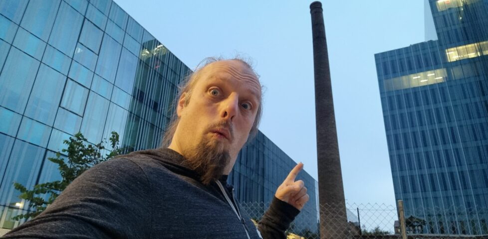 Dan points at an old brick chimney, standing alone among modern glass office buildings.