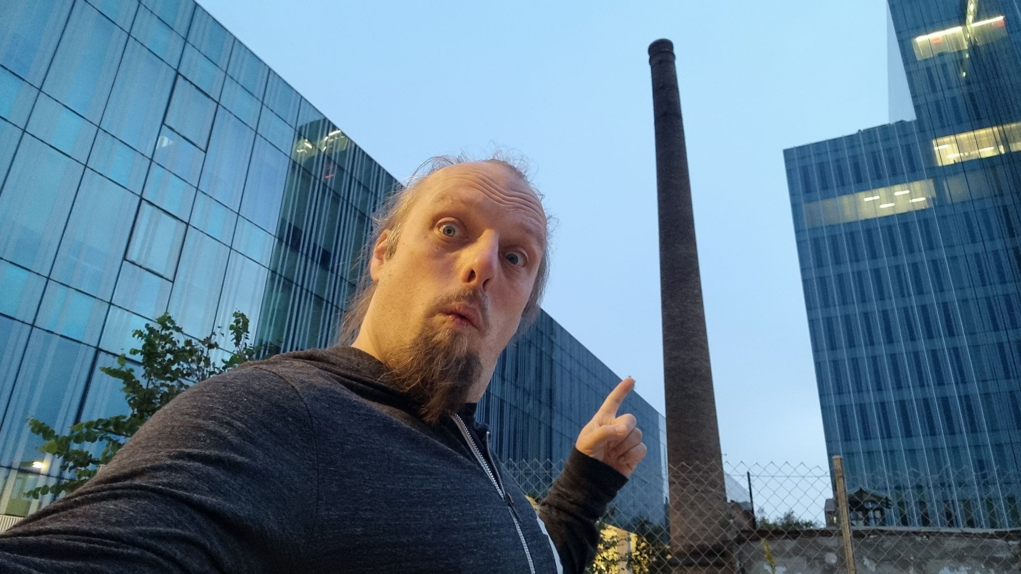 Full image of Dan points at an old brick chimney, standing alone among modern glass office buildings.