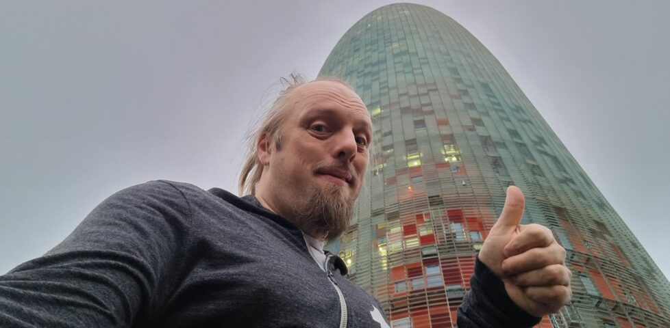 Under a cloudy early-morning sky, Dan gives a thumbs-up in front of a gherkin-shaped skyscraper.