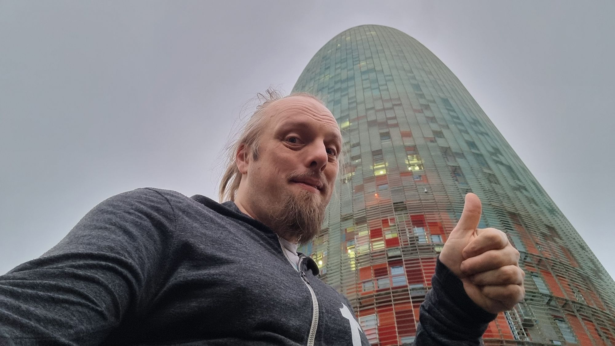 Full image of Under a cloudy early-morning sky, Dan gives a thumbs-up in front of a gherkin-shaped skyscraper.