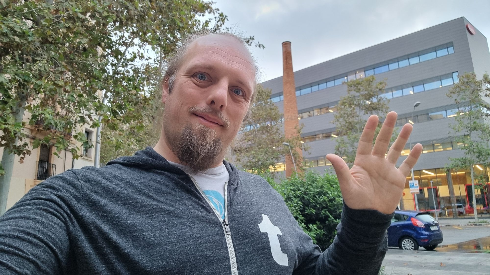 Full image of Dan waves in front of an old brick chimney surrounded by modern office and retail buildings.