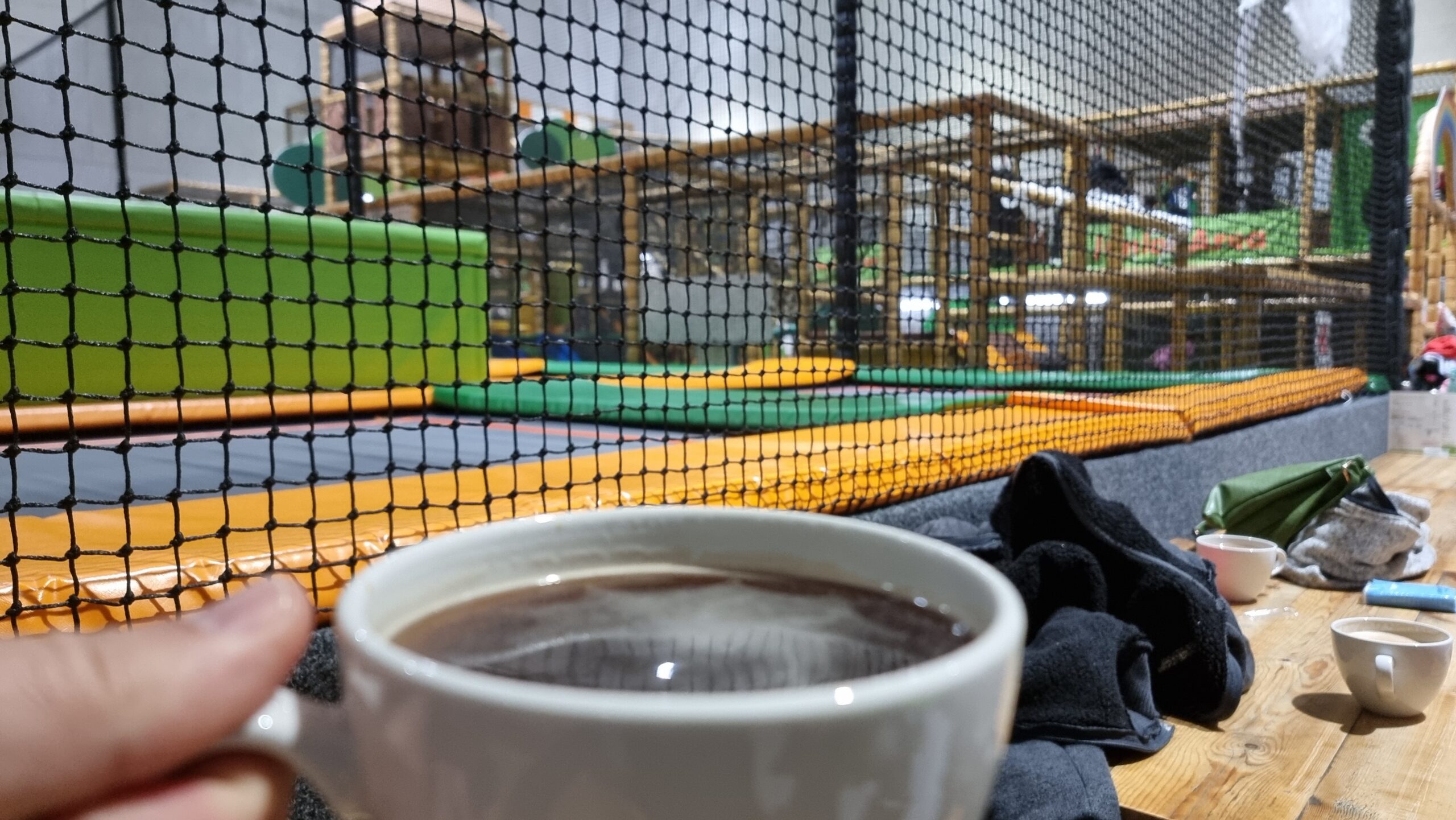 Full image of A mug of coffee held in front of a view of a multicoloured soft playground.
