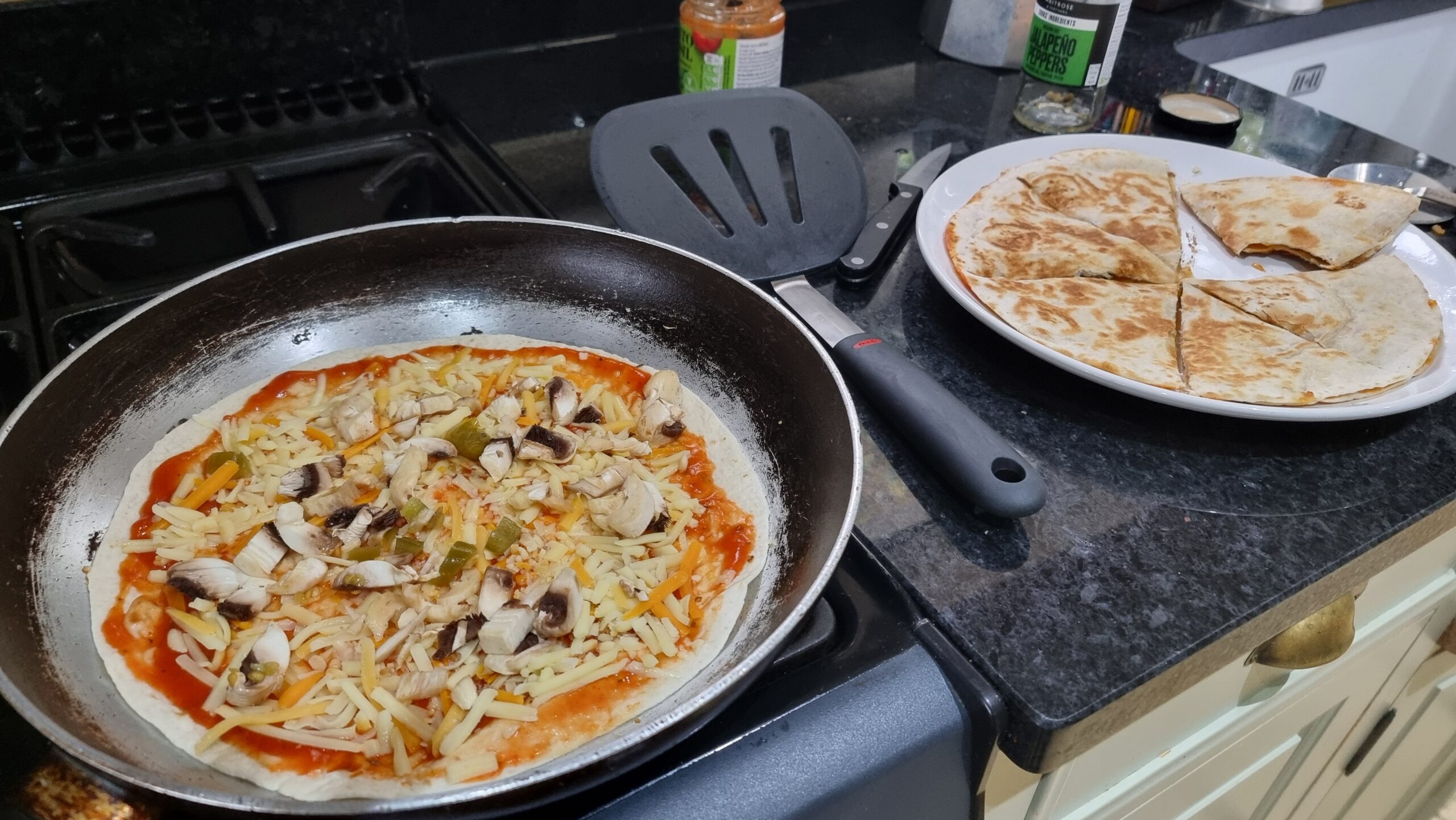 Full image of Gas stovetop.a frying pan contains a tortilla wrap topped with tomato sauce, cheese, mushrooms, and jalapeños. Beside its a plate containing a completed quesapizza: two crispy tortilla wraps sandwiching their contents.