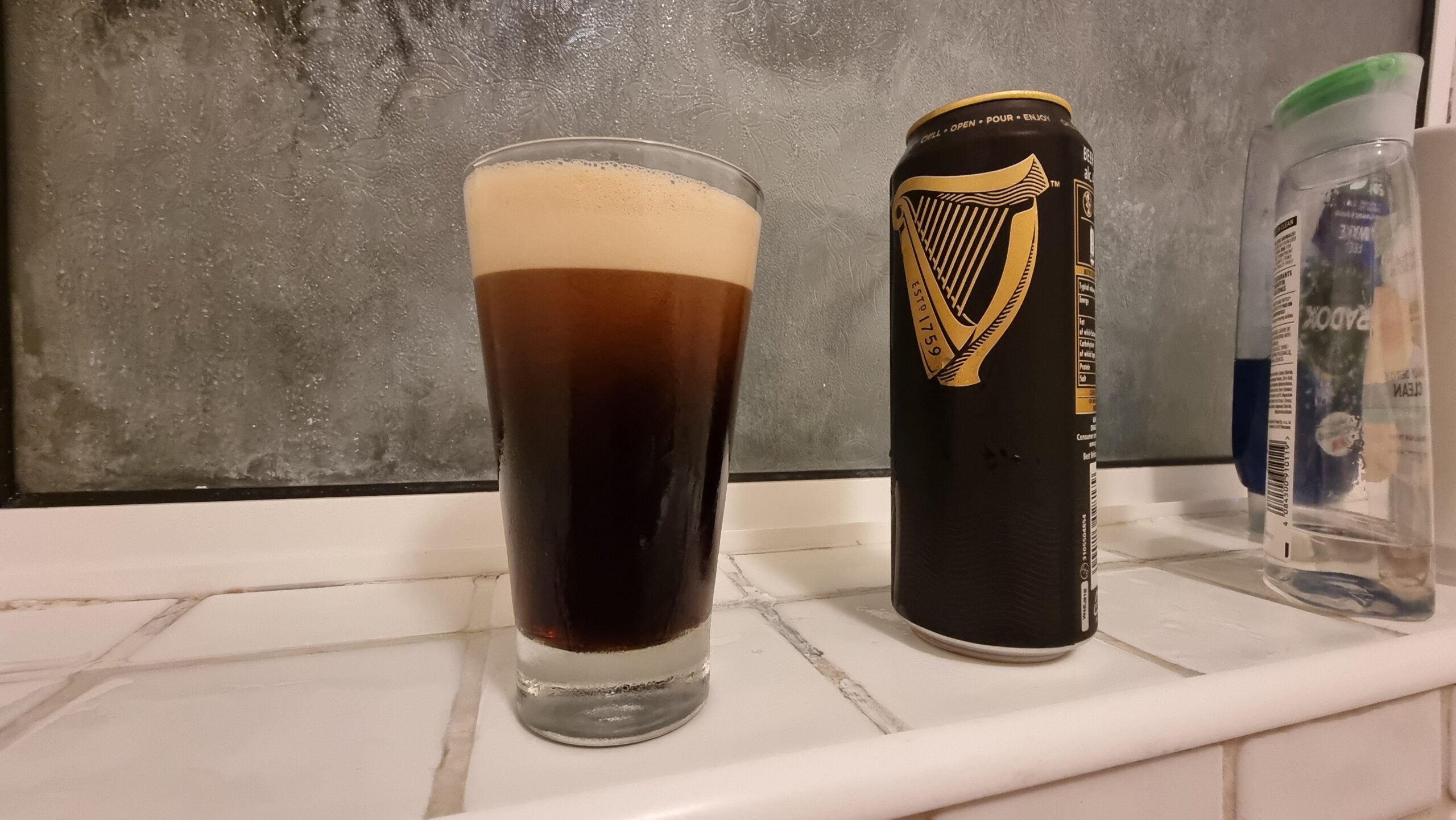 Full image of A pint of Guinness alongside a can, on a tiled bathroom shelf.