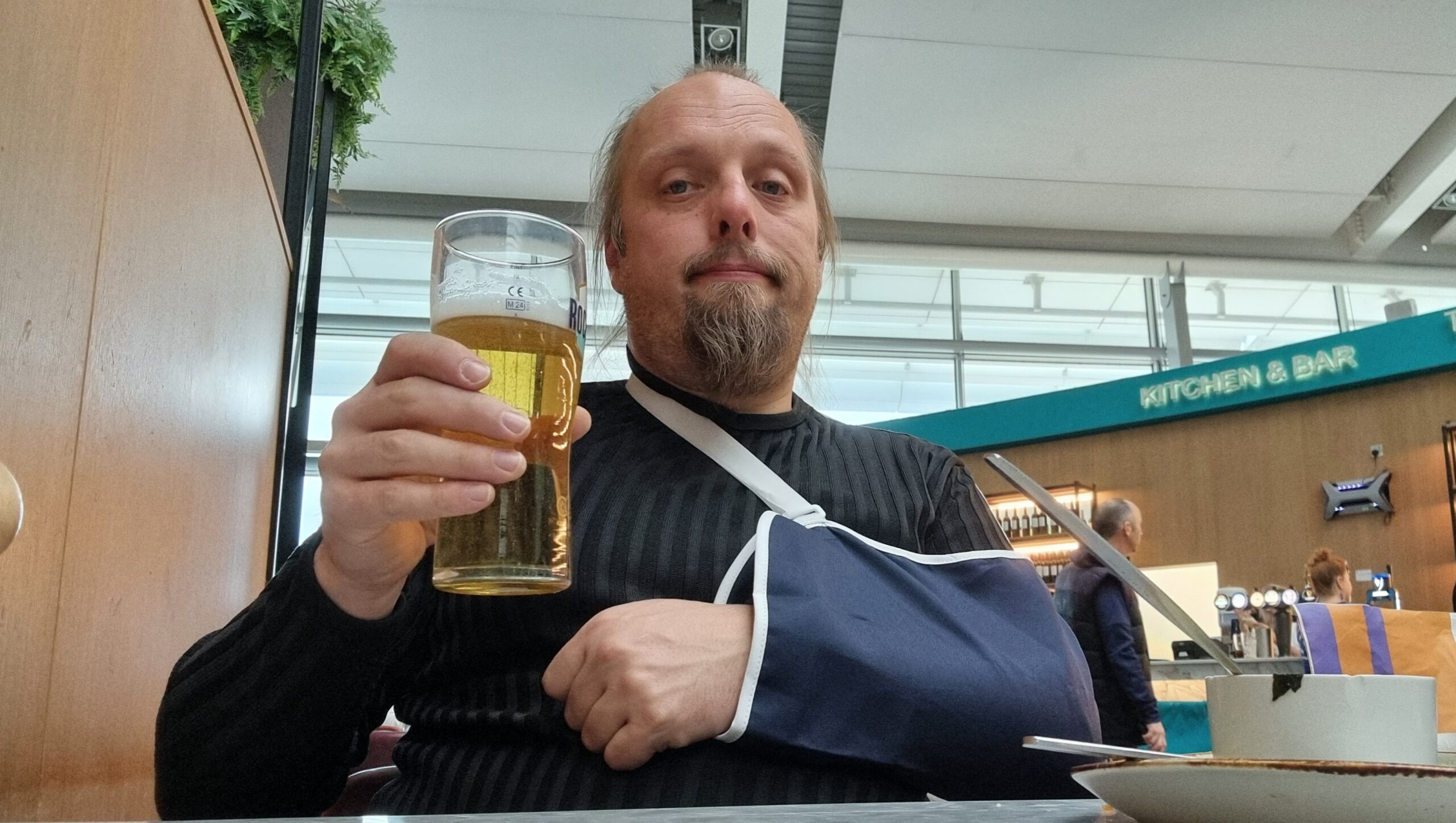Full image of Dan sits in an airport bar drinking a beer held in his right hand. His left arm is in a sling.