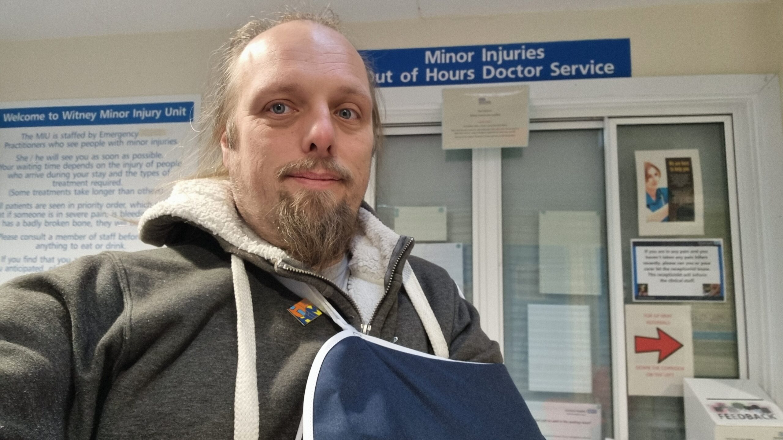 Full image of Dan, wearing a sling, stands near the reception of a Minor Injuries Unit.