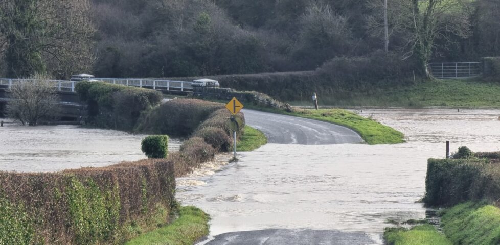 A road through a field and towards a river bridge is deeply submerged under fast-flowing water.
