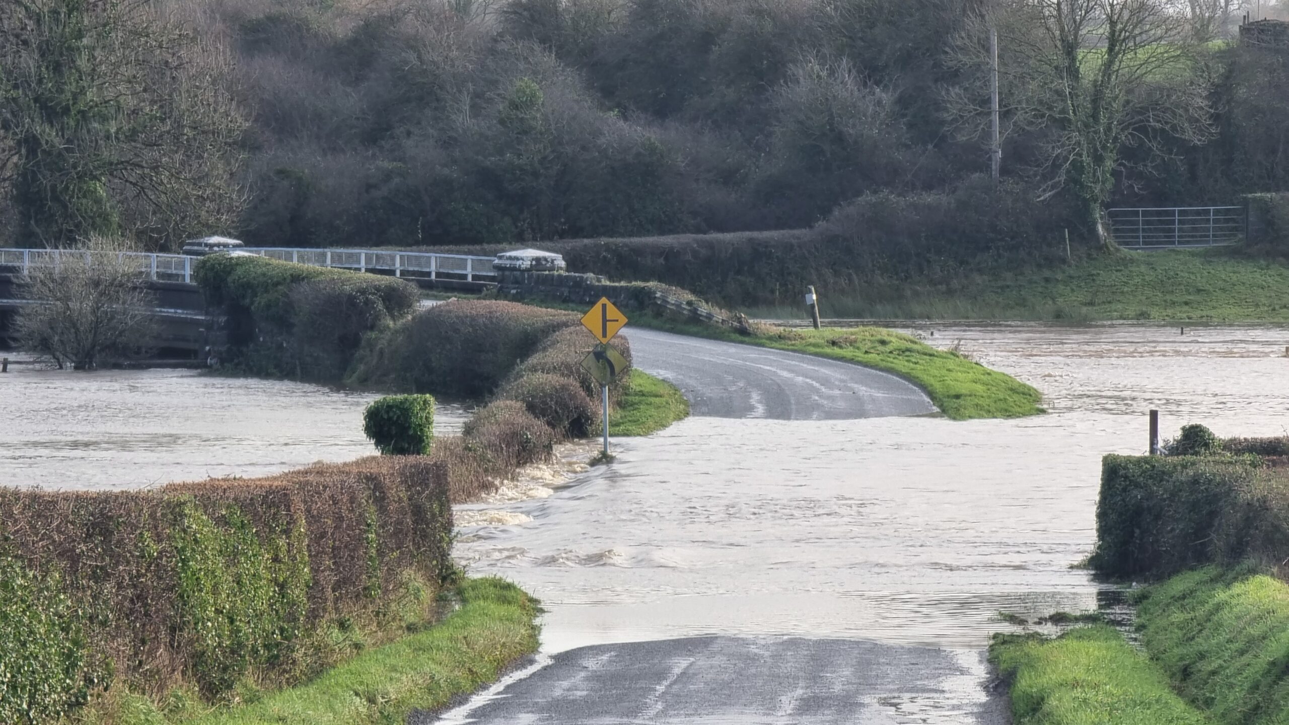 Full image of A road through a field and towards a river bridge is deeply submerged under fast-flowing water.