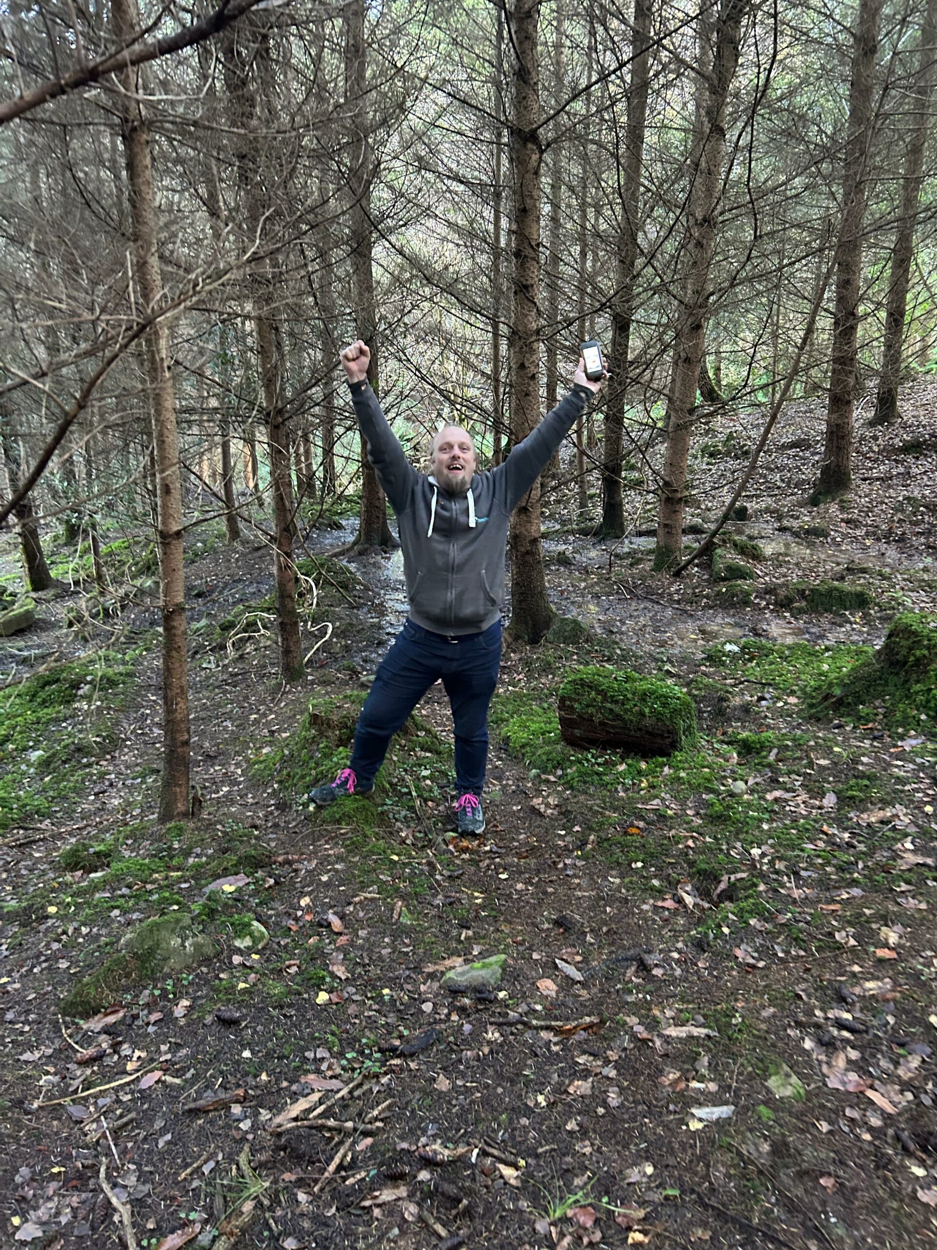 Full image of Dan raises his hands in a victory pose in a forest.