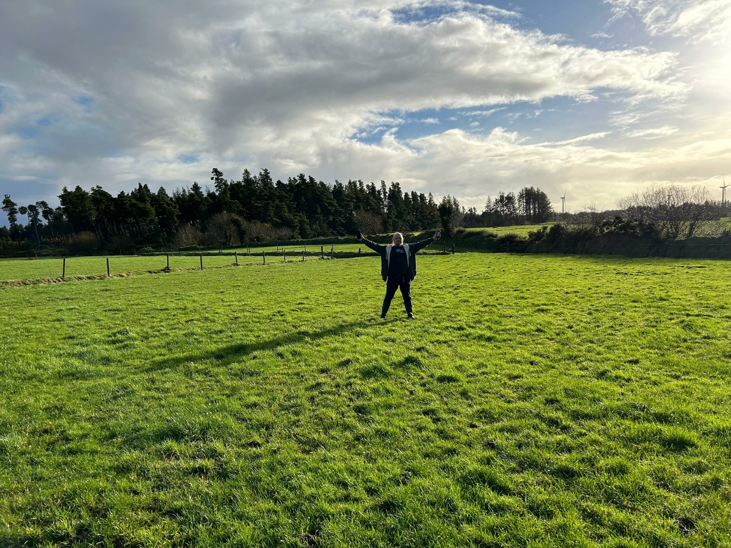 Full image of Dan raises his hands - one of them holding a GPS receiver - in the air in a victory pose, in a green pasture with a forest in the background.