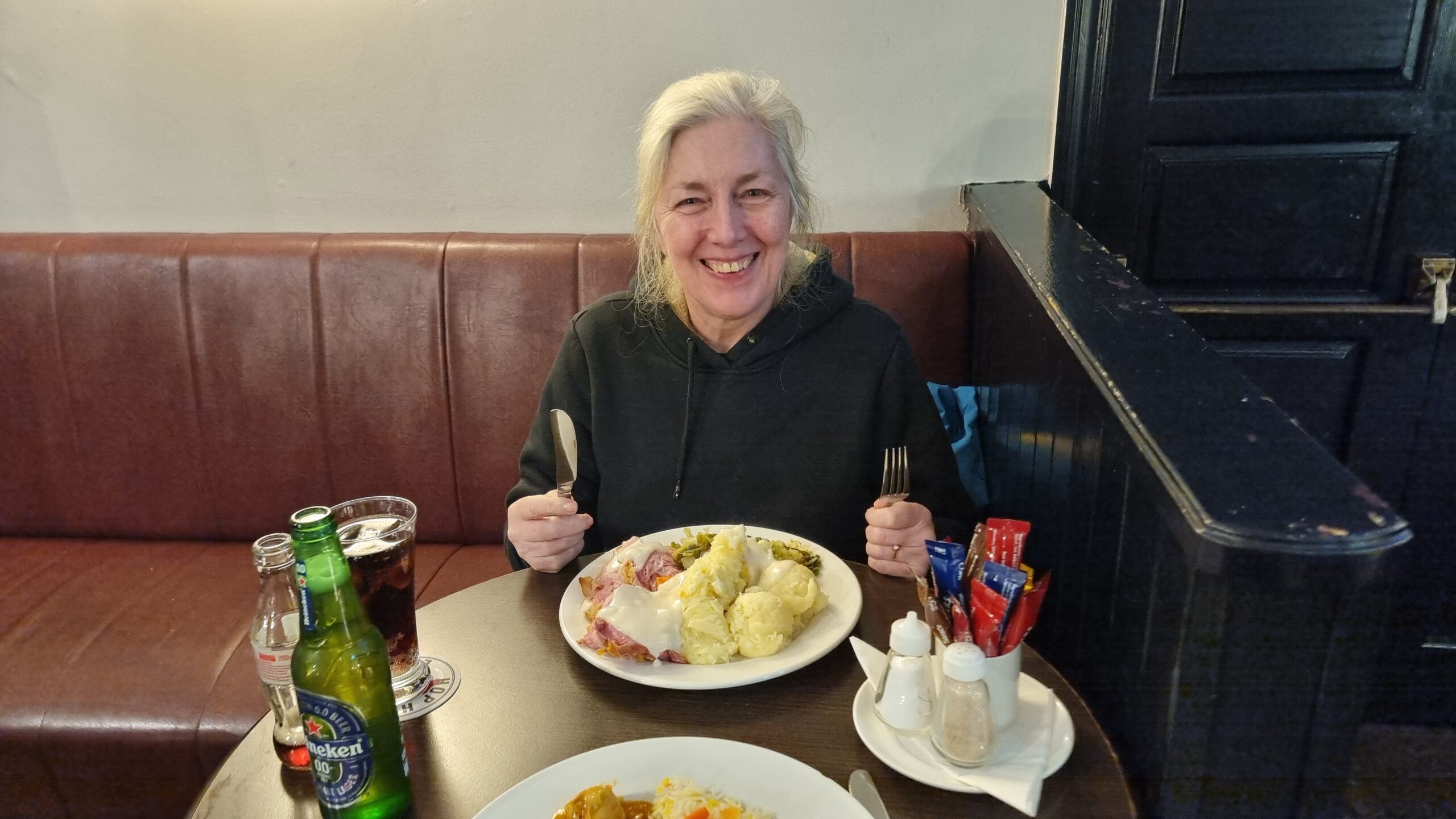 Full image of A woman sits in front of a plate loaded with food in a pub setting, holding a knife and fork.