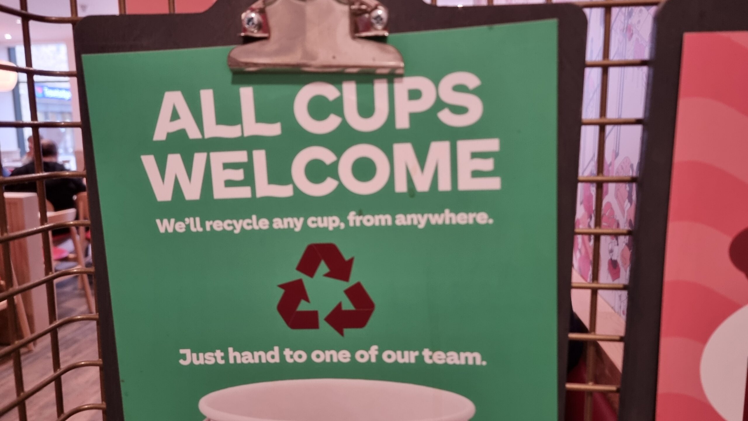 Full image of Sign made from a clipboard, in a coffee shop, reading "All cups welcome. We'll recycle any cup, from anywhere. Just hand to one of our team."
