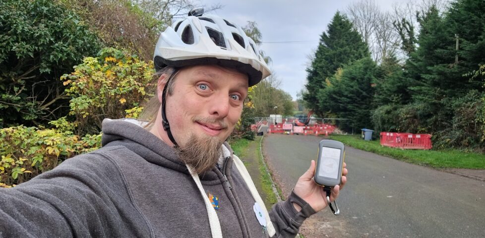 Dan smiling, wearing a cycle helmet, holding a GPSr by the side of a narrow road, with roadworks in the background.