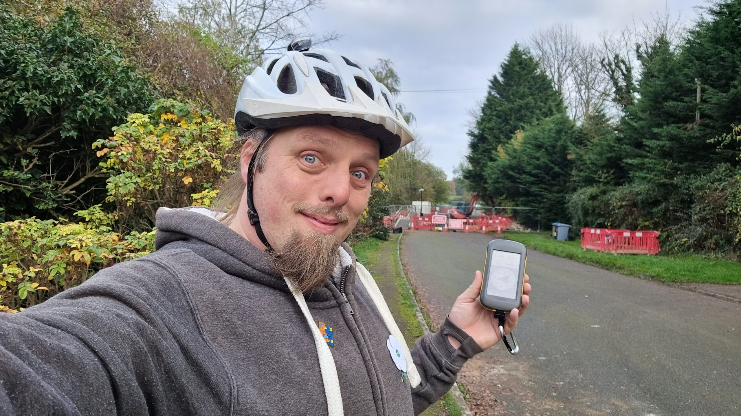 Full image of Dan smiling, wearing a cycle helmet, holding a GPSr by the side of a narrow road, with roadworks in the background.