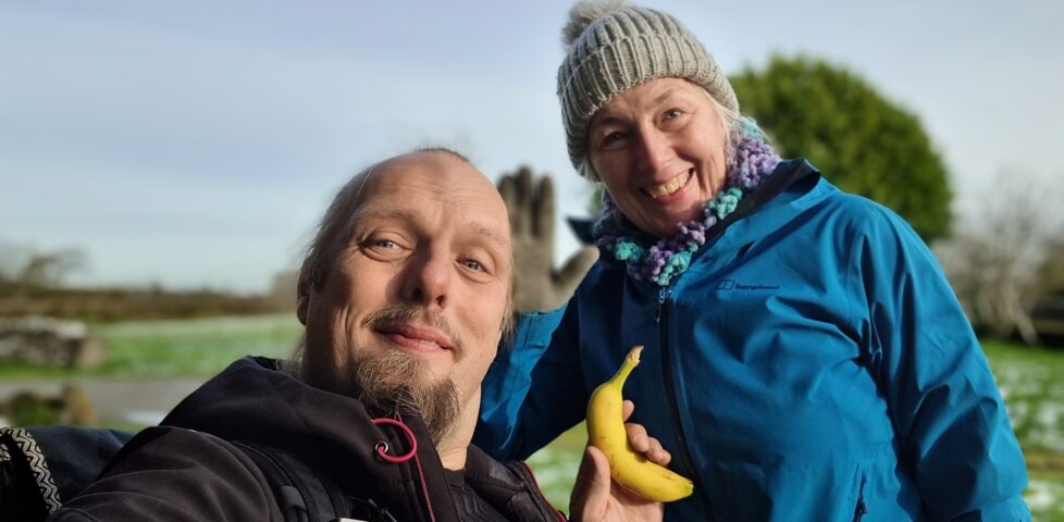 Dan and his mother smiling in a field. Dan is holding a banana.