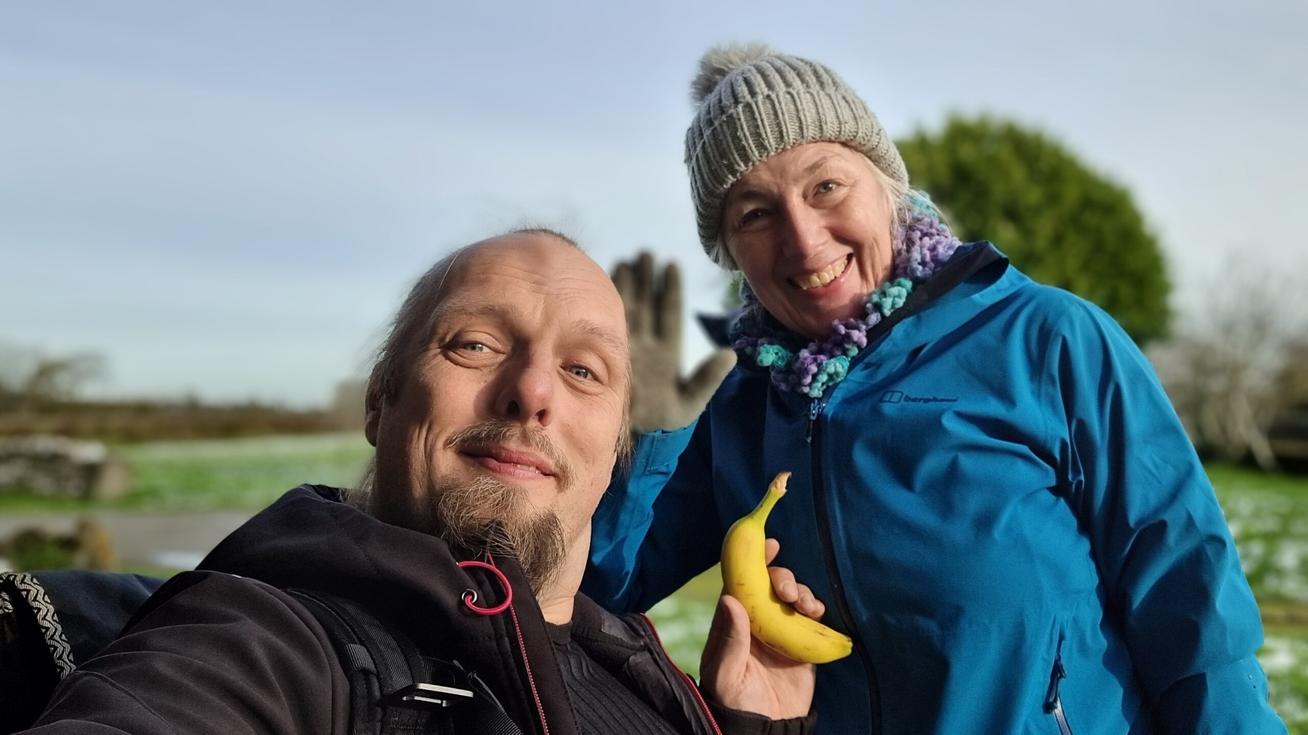 Full image of Dan and his mother smiling in a field. Dan is holding a banana.