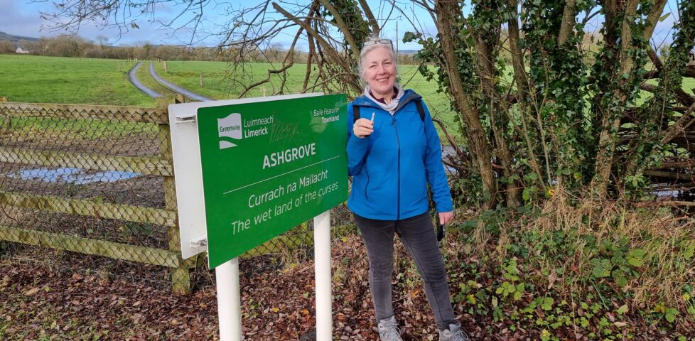 A smiling woman with white hair, wearing spectacles atop her head, stands next to a sign that reads 'Ashgrove: The wet land of the curses' in English and Irish. She holds a small tube-shaped geocache.