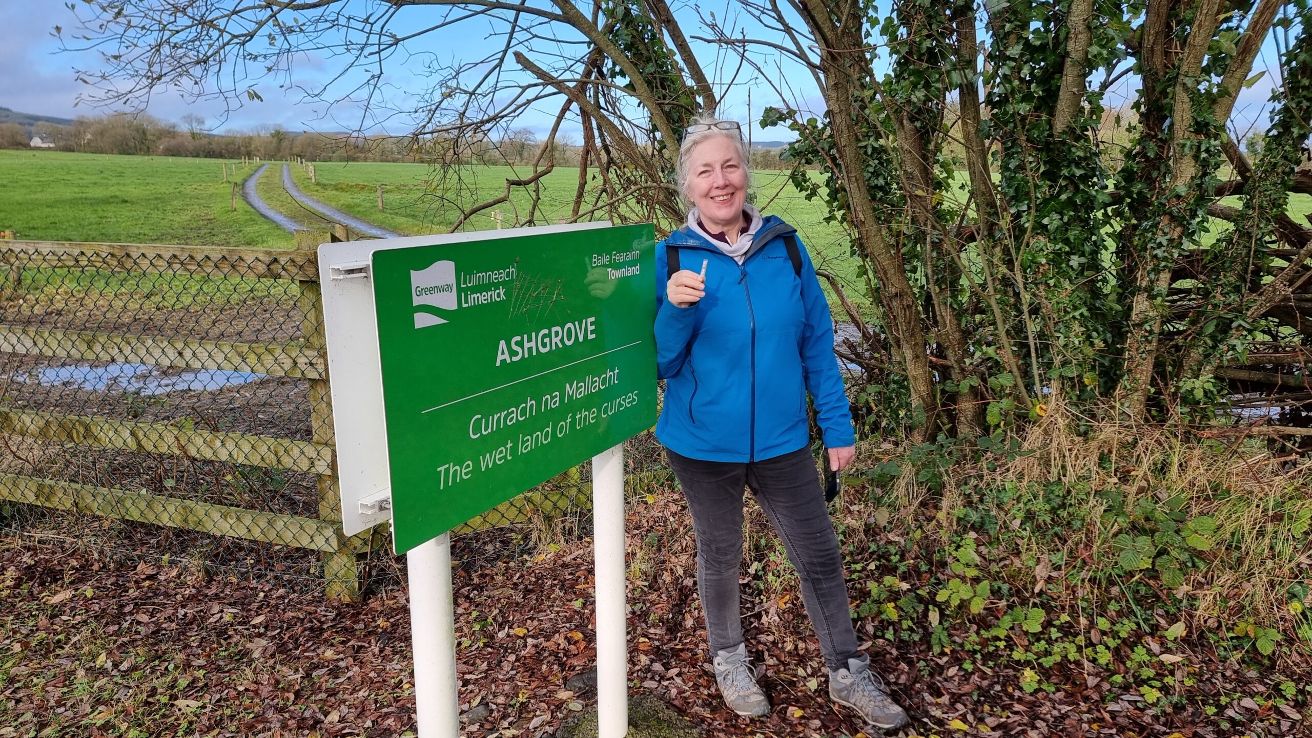 Full image of A smiling woman with white hair, wearing spectacles atop her head, stands next to a sign that reads 'Ashgrove: The wet land of the curses' in English and Irish. She holds a small tube-shaped geocache.