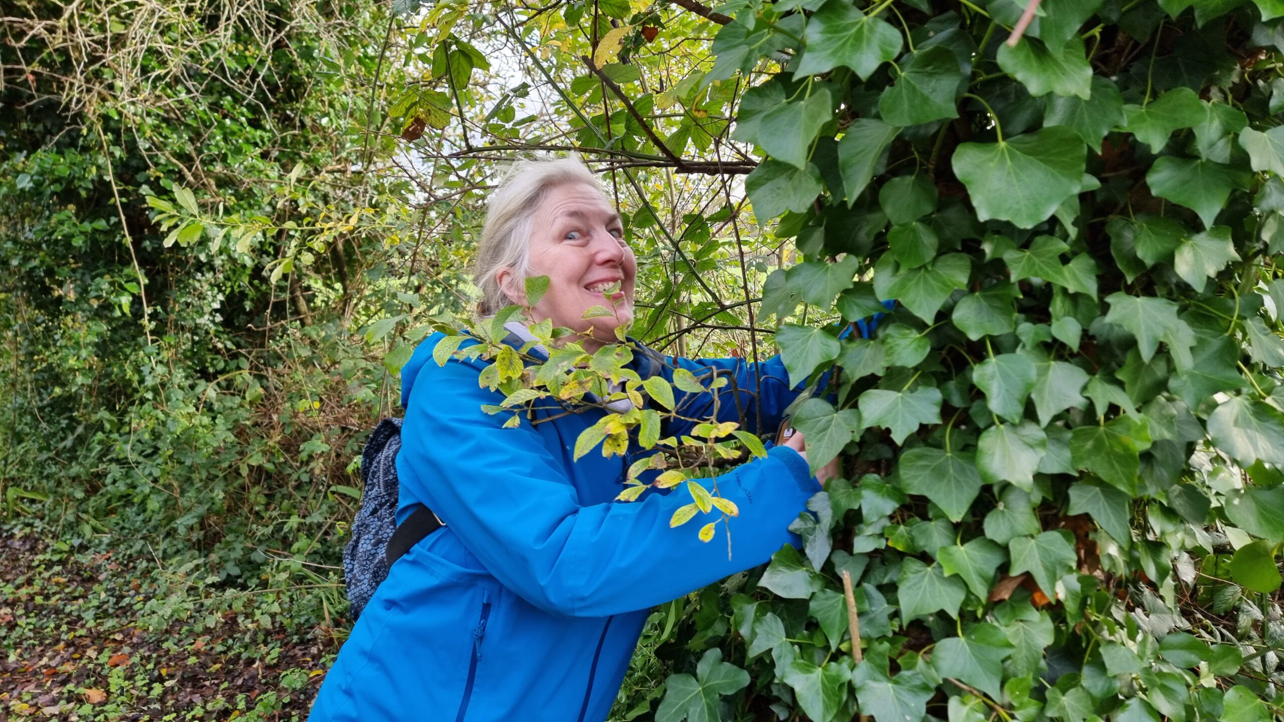 Full image of A woman with white hair, wearing a blue coat, grins maniacally as she reaches deep into an ivy-covered tree.