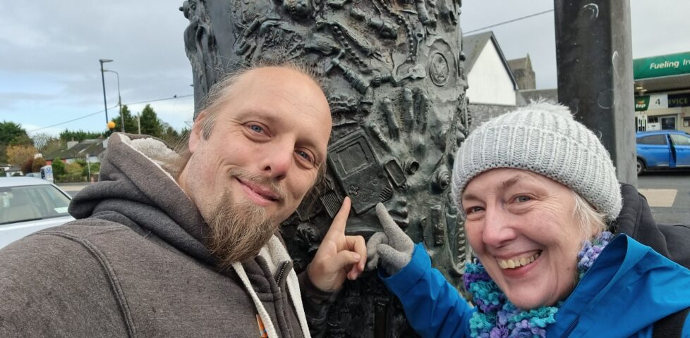 A cylindrical metal sculpture stands upright on a spindle, in a manner slightly reminiscient of a kebab. It's decorated with hundreds of small contemporary handheld objects such as spanners, rings, spoons, and chess pieces. Dan and his mother Doreen stand in front, both pointing to a Nintendo Game Boy.