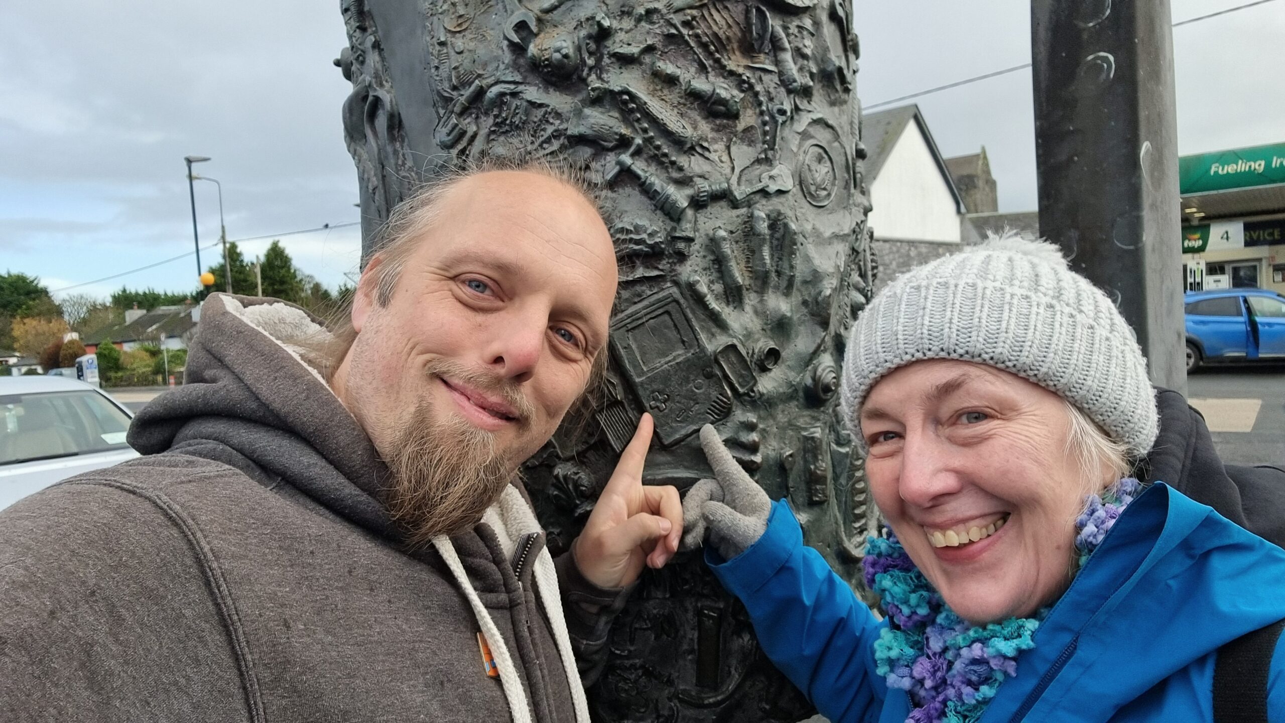 Full image of A cylindrical metal sculpture stands upright on a spindle, in a manner slightly reminiscient of a kebab. It's decorated with hundreds of small contemporary handheld objects such as spanners, rings, spoons, and chess pieces. Dan and his mother Doreen stand in front, both pointing to a Nintendo Game Boy.