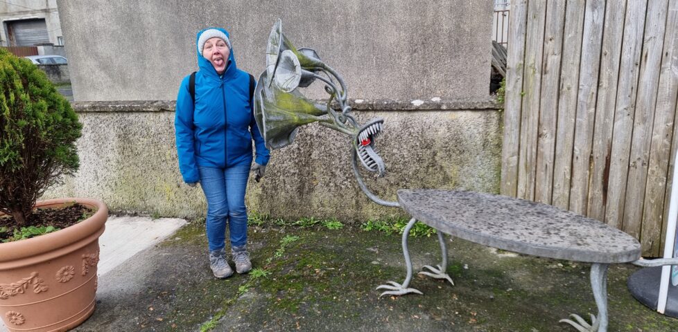 A sculpture (called 'gossip') is a bench with birdlike feet and a head comprised of a mouth with a long tongue and several listening tubes. Alongside it, Dan's mother - a woman wearing a grey bobble hat and a blue raincoat - attempts to mime the same mouth shape.