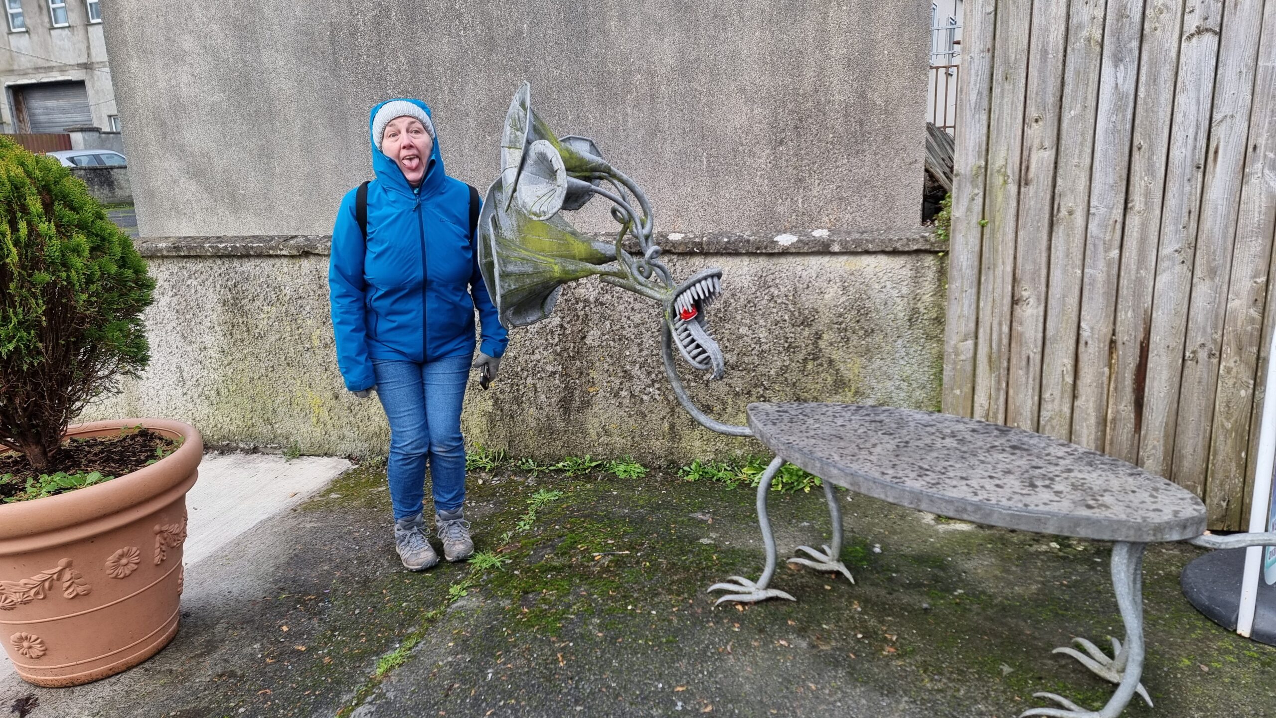 Full image of A sculpture (called 'gossip') is a bench with birdlike feet and a head comprised of a mouth with a long tongue and several listening tubes. Alongside it, Dan's mother - a woman wearing a grey bobble hat and a blue raincoat - attempts to mime the same mouth shape.