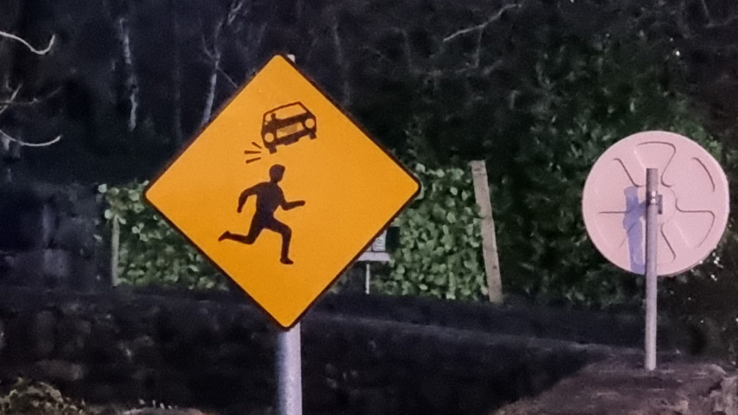Full image of Photograph showing a road sign; it's yellow, diamond-shaped, and depicts in black the silhouette of a person running from left to right. Above them is the silhouette of a car, much smaller than them and twisted anticlockwise by about 20 degrees. Impact/movement marks eminate from the lower of the car's wheels, as if it's a thrown object that's just bounced off the head of the runner.