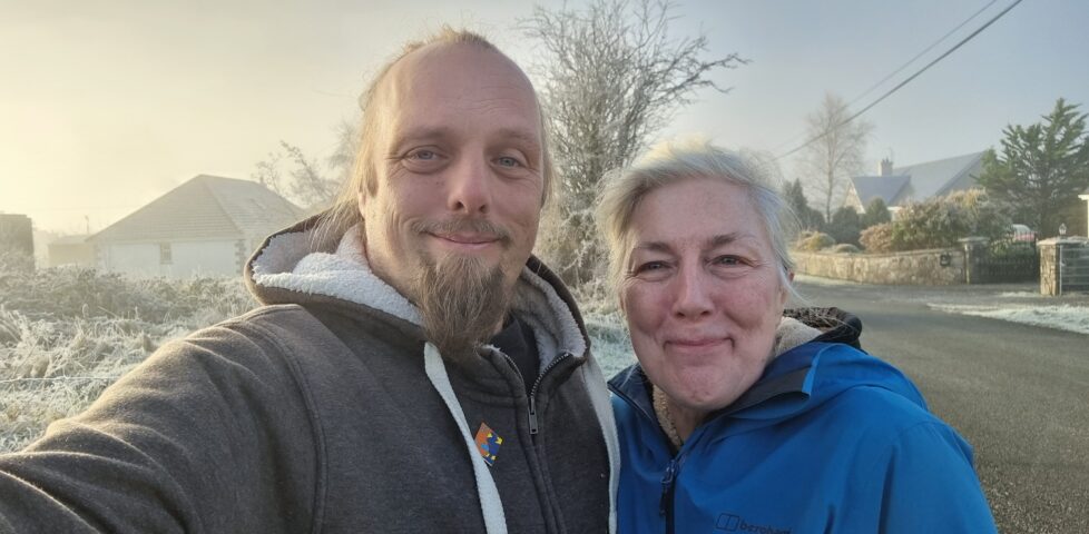 Dan and his mother, smiling, in frozen countryside, with fog behind.