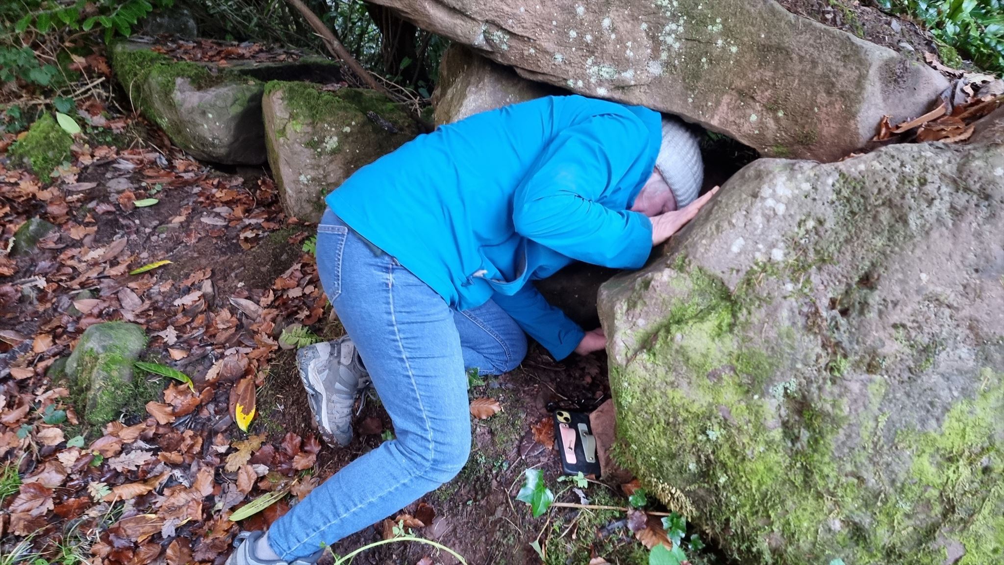 Full image of A woman in a bobble hat and a blue raincoat crawls into a gap between two boulders.