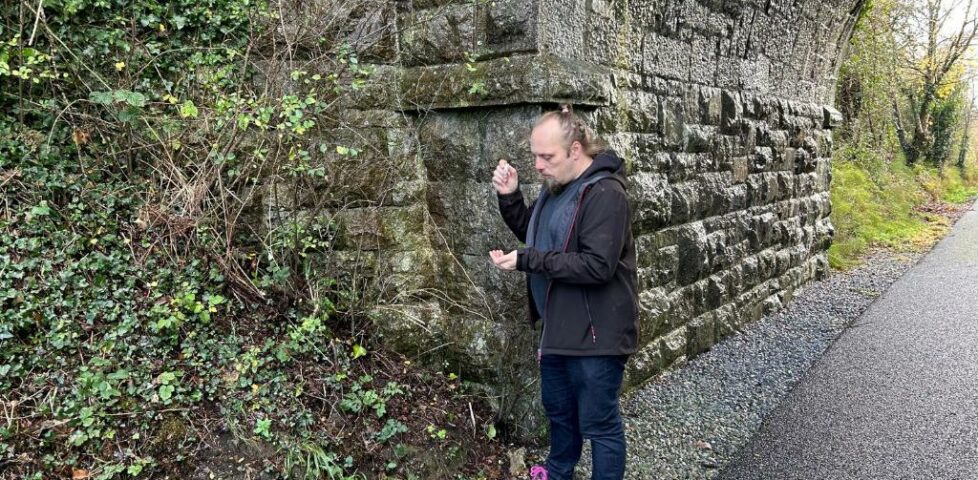 Dan, standing by a bridge, shakes a logbook out of a tubular geocache.