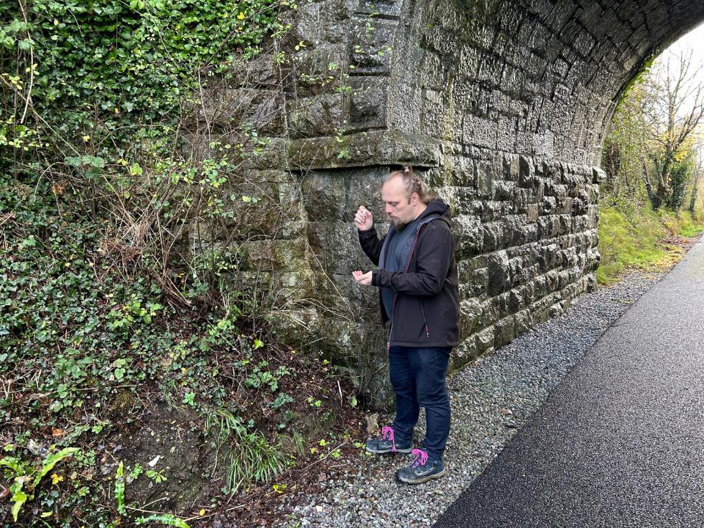 Full image of Dan, standing by a bridge, shakes a logbook out of a tubular geocache.