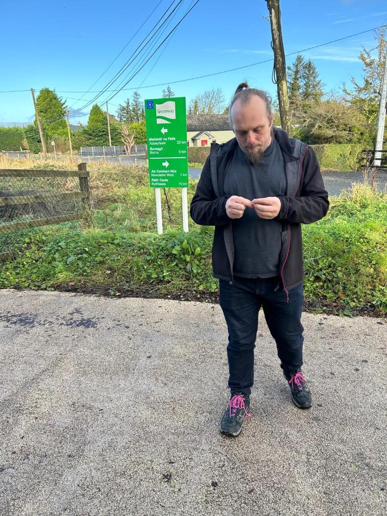 Full image of Dan, standing by a "Greenway" sign, unrolls a geocaching logbook.