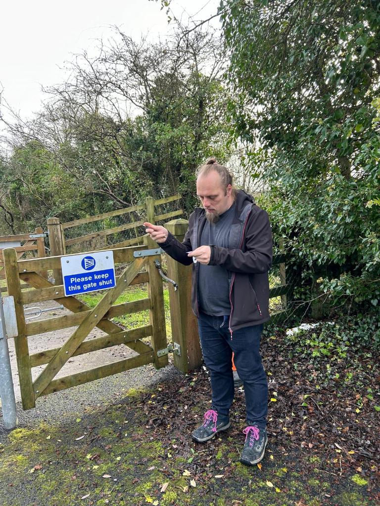 Full image of Dan standing by a gate, reading a geocaching logbook.