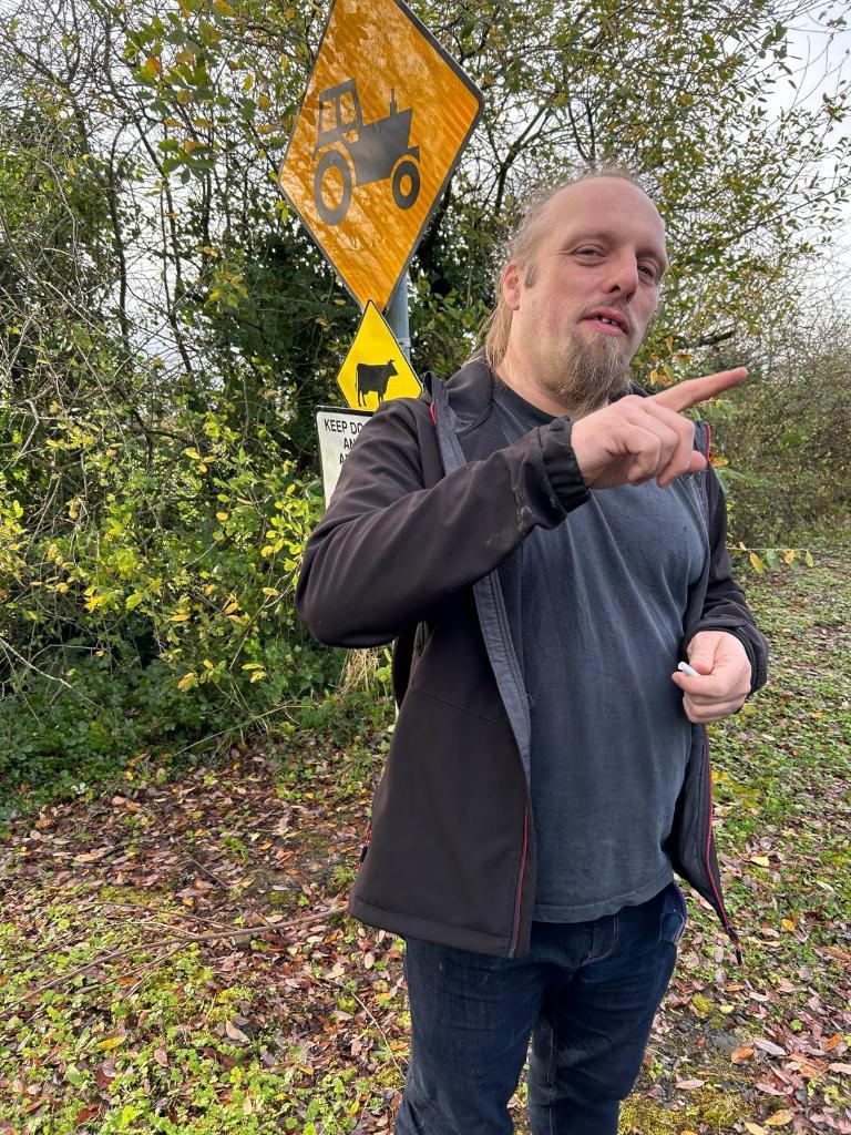 Full image of Dan, with his finger sticking out, stands in front of 'cow crossing' and 'tractor crossing' signs.