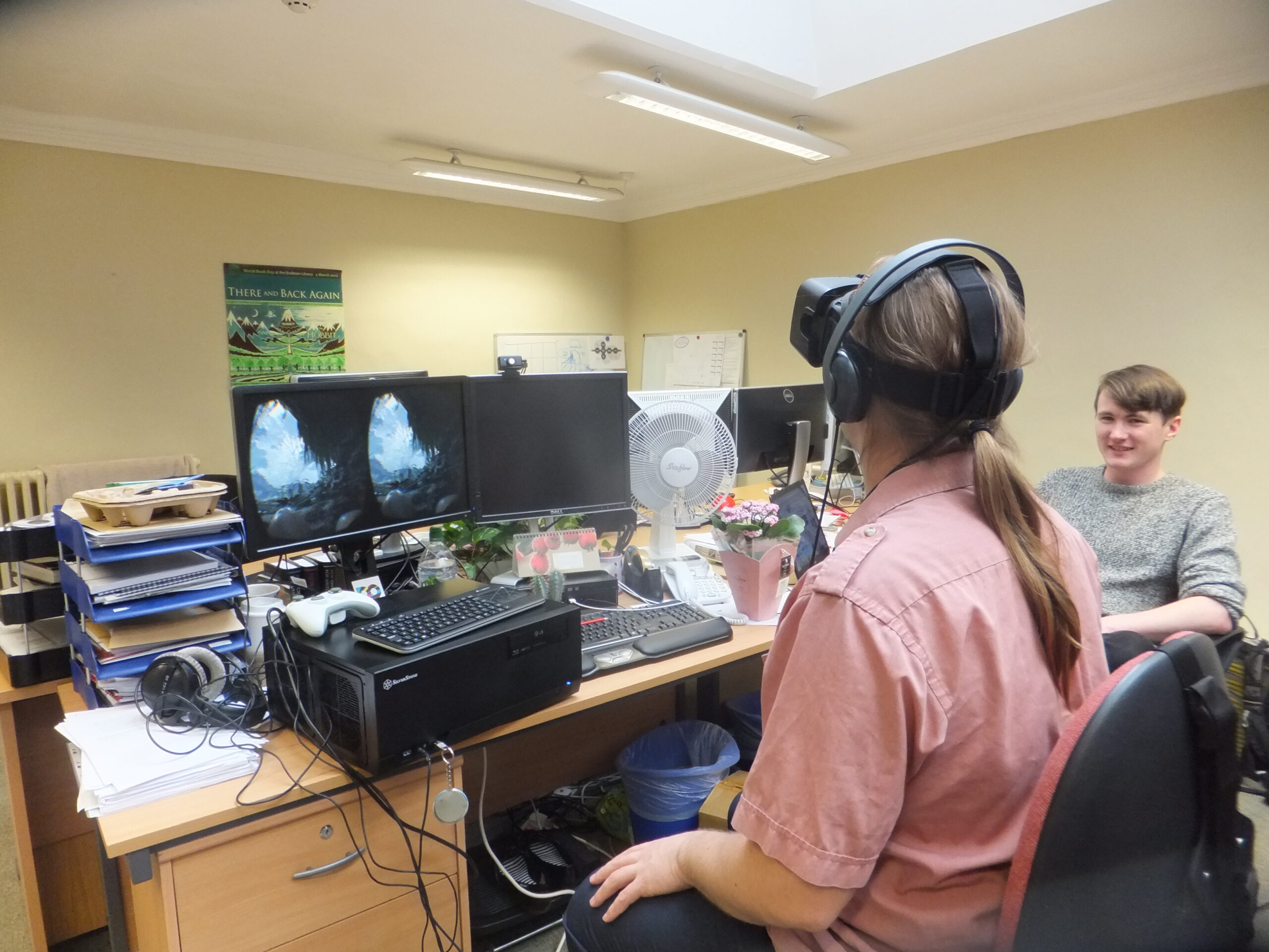 Full image of Dan, wearing a VR headset, sits in an office environment, watched by Adam.