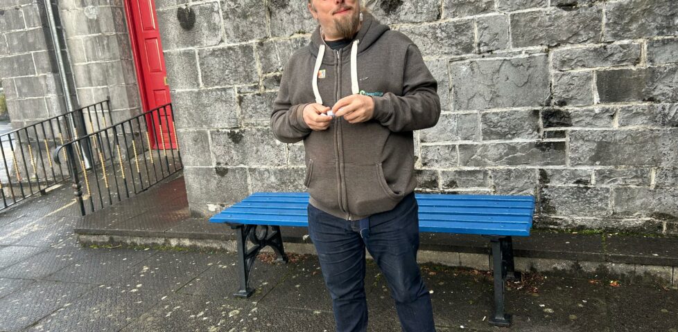 Dan stands in front of a blue bench, in the rain, in front of a stone wall.