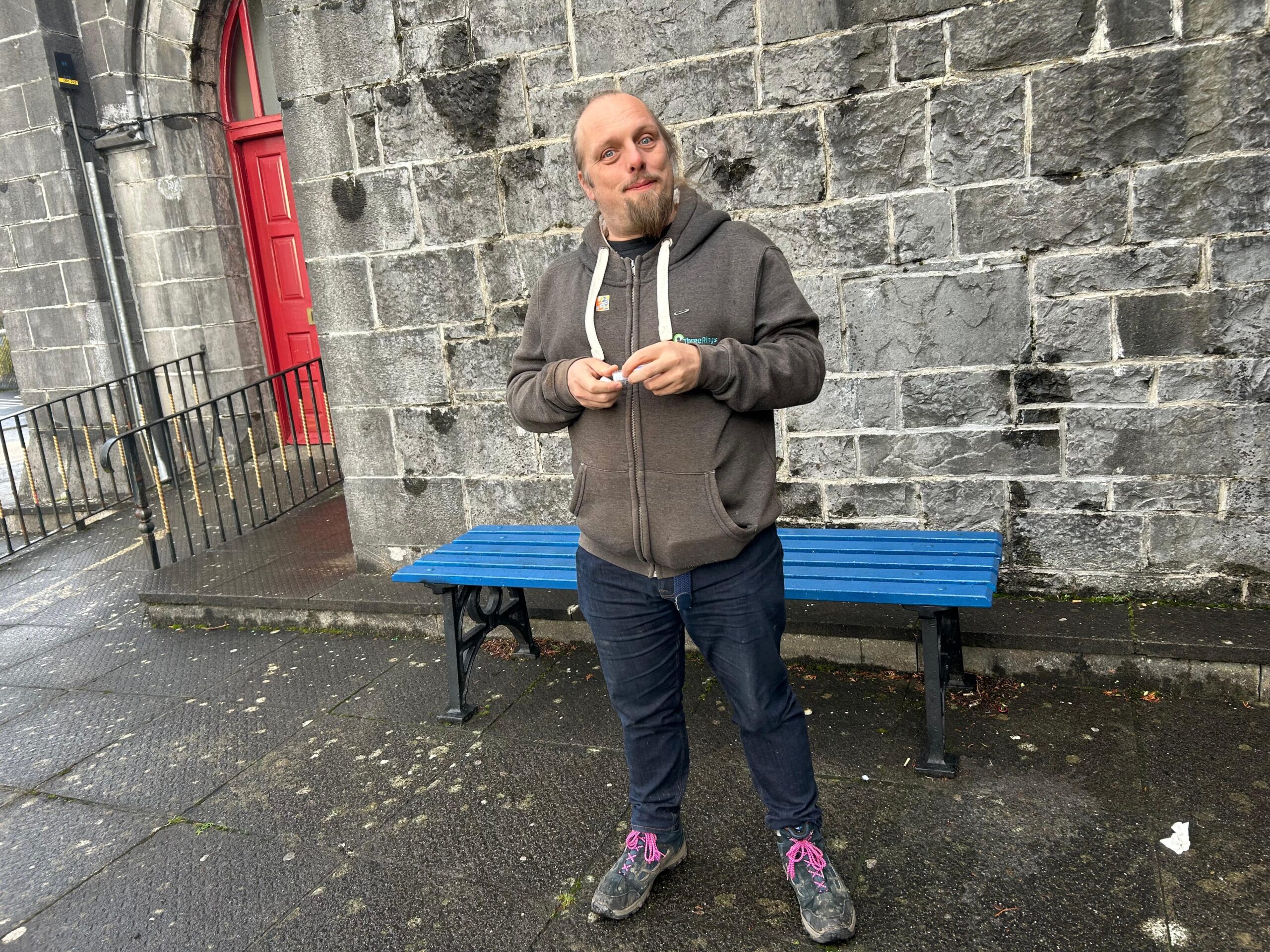 Full image of Dan stands in front of a blue bench, in the rain, in front of a stone wall.