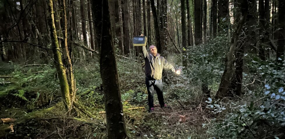 Dan, in a dense forest at twilight, holds aloft a large ammo can. His other hand holds his phone, used as a flashlight.