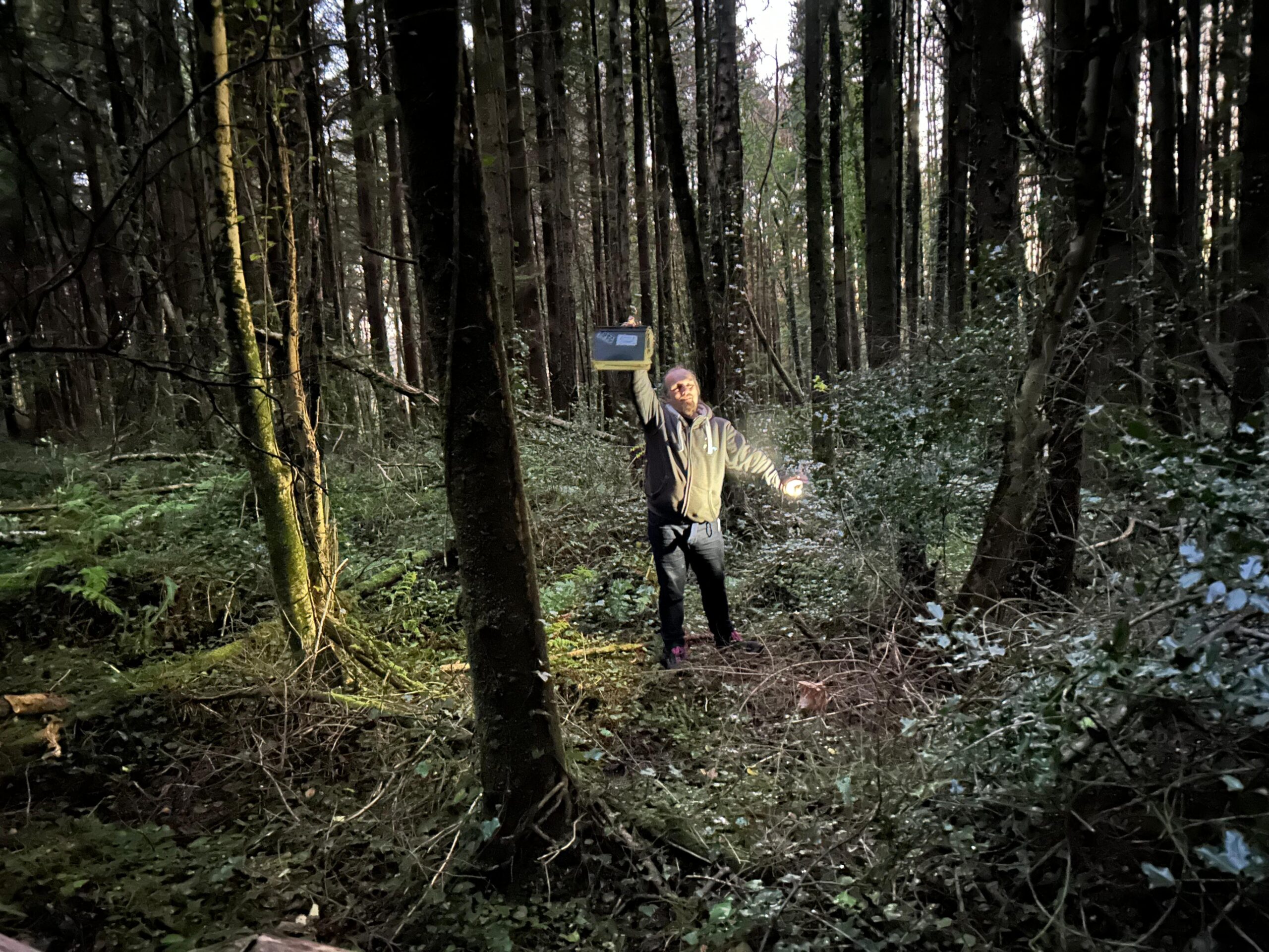 Full image of Dan, in a dense forest at twilight, holds aloft a large ammo can. His other hand holds his phone, used as a flashlight.
